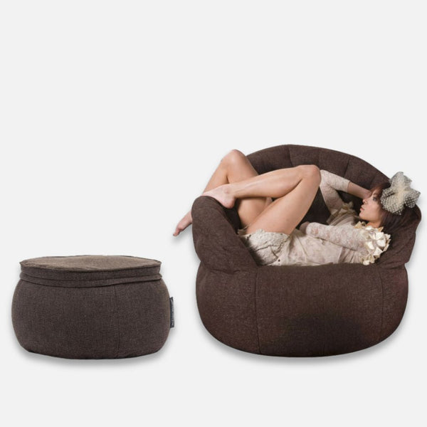 Kona í léttum fötum og með hatt slakar á í stórum, kringlóttum Butterfly Chaise hægindastól. Við hlið hennar er samsvarandi minnisfroðubekkur, báðir með mjúku og þægilegu útliti. Bakgrunnurinn er hvítur.