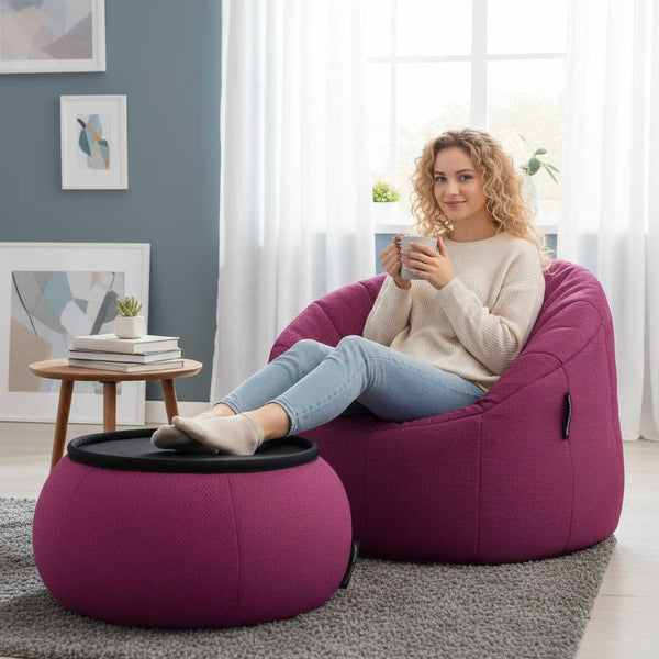 Kona með ljóst, krullað hár situr á Ambient Lounge Cove Package-baunasekk, heldur á bolla og hvílir fæturna á samsvarandi fótskör í notalegri stofu fullri af úramjúku efni og nútímalegum, þægilegum húsgögnum.