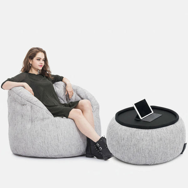 Kona í dökkum kjól slakar á í ljósgráum baunasekk frá Ambient Lounge, með fæturna á samsvarandi fótlegg með svörtum bakka sem heldur spjaldtölvu. Þægilega húsgagnasettið stendur á hvítum bakgrunni.
