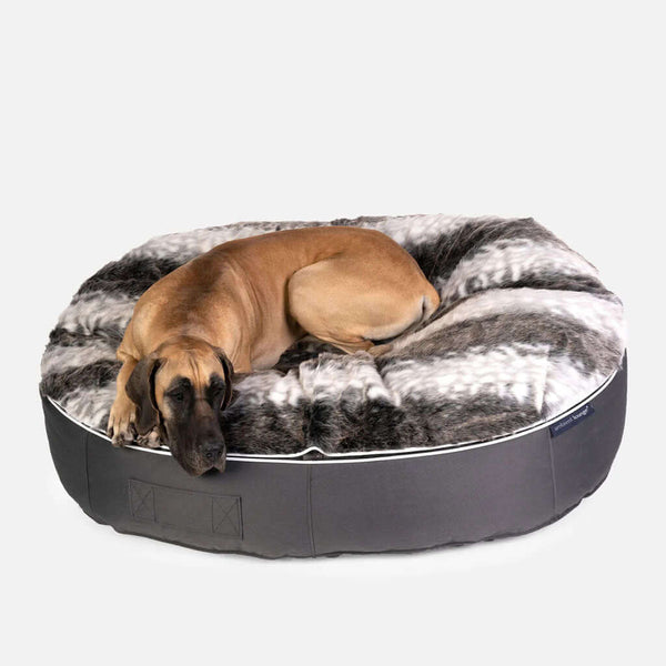 Stór brúnn hundur liggur á Ambient Lounge hundeseng XL, kringlóttu, mjúku hundarúmi með gráum og hvítum gervifeldstoppi og dökkgráum botni úr efni, á hvítum bakgrunni.