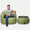 Brosandi maður í gráum stuttermabol og bláum gallabuxum situr í grænum Ambient Lounge® baunastól við hliðina á samsvarandi grænum fótskör á einföldum ljósum bakgrunni.